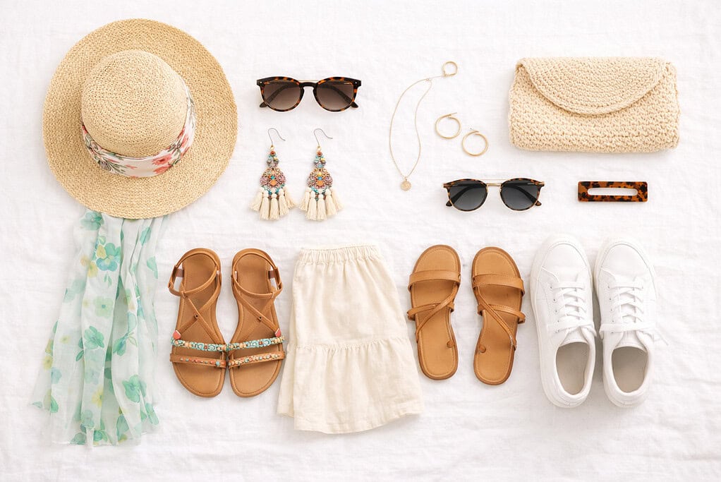 Flat lay of summer accessories and shoes including a floral straw hat, boho earrings, sunglasses, gold jewelry, sandals, white sneakers, a scarf, and a woven clutch on a white fabric background.