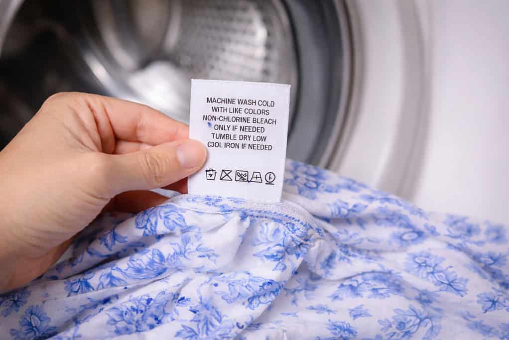 Close-up of a hand holding a clothing care label with washing instructions, on a neutral flat-lay background.