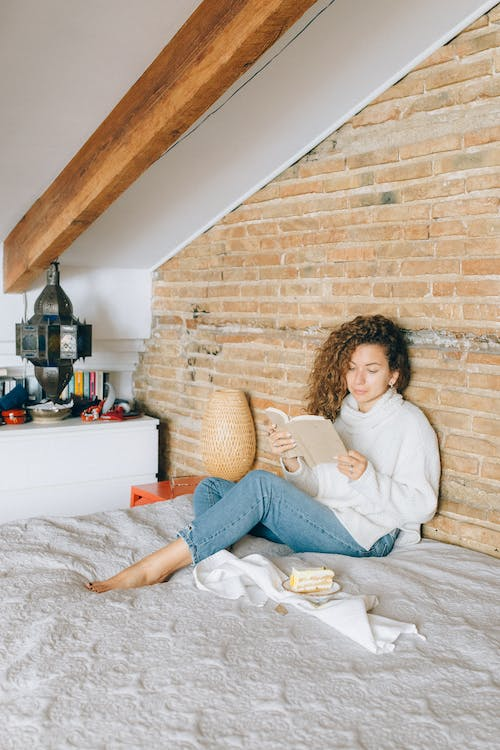 cozy woman in white sweater reading a book with sandwich beside her.