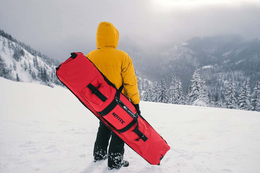 a person standing on a snowy mountain slope, viewed from behind, holding a long red snowboard bag secured with black straps