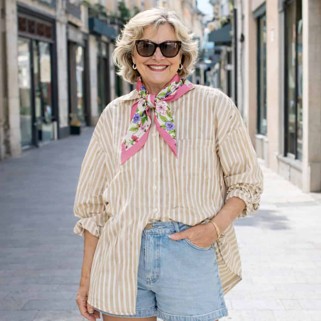 A mature woman wearing sunglasses, a striped shirt with rolled-up sleeves, denim shorts, and a pink floral scarf poses confidently on a sunlit city street.