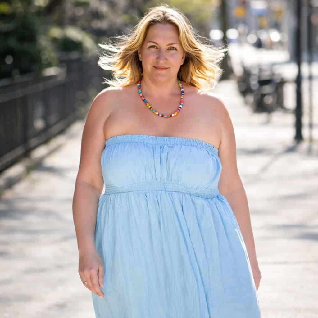 A smiling blonde woman in a light blue strapless dress and colorful beaded necklace walks outside on a sunny day.