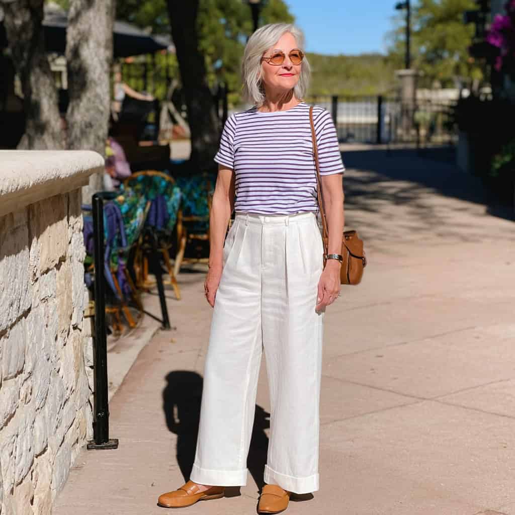 Chic older woman in a classic Breton striped tee paired with high-waisted white wide-leg trousers, tan loafers, and sunglasses for a timeless Parisian-inspired look.