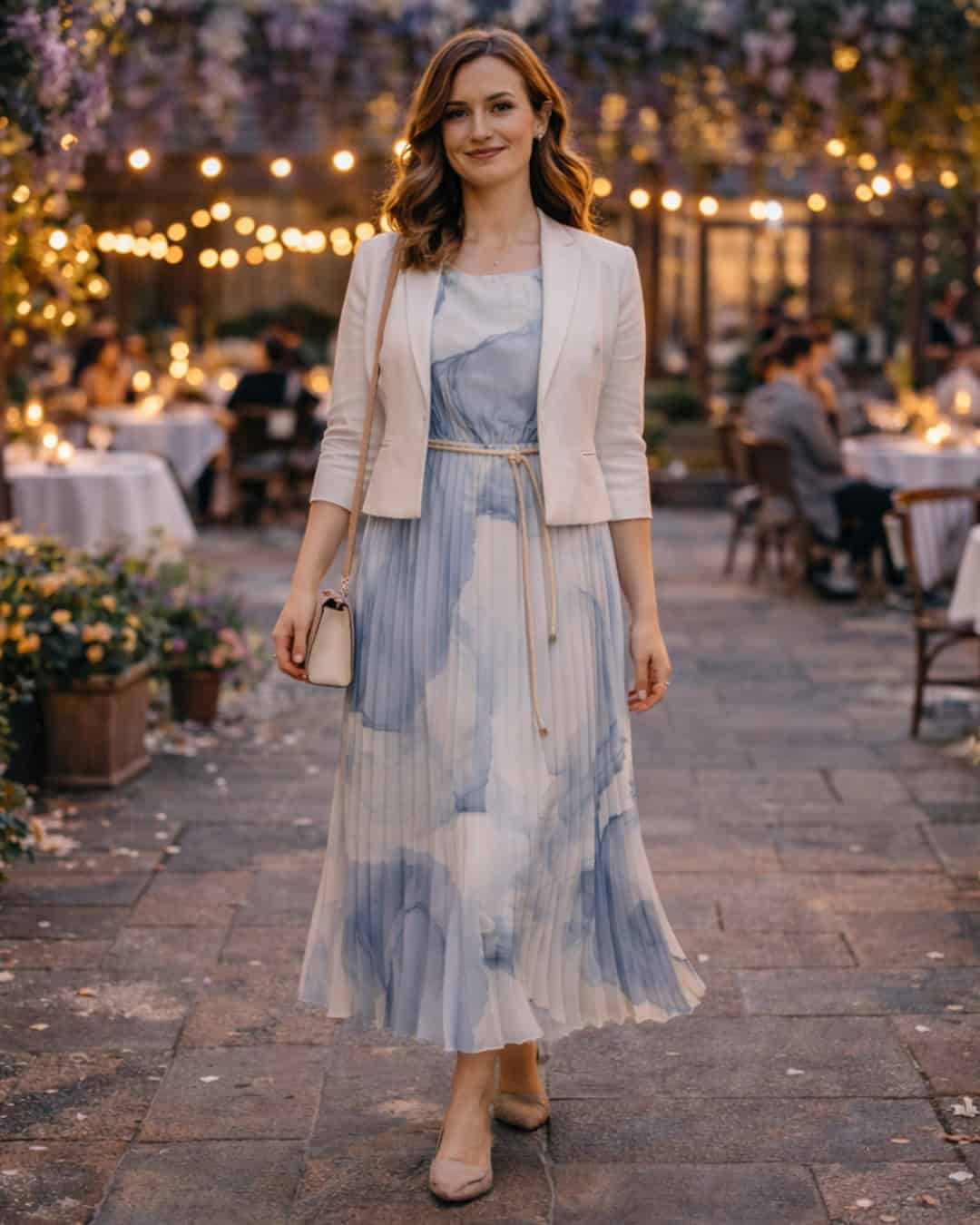 Woman in a blue-and-white pleated midi dress with a light blazer walking through a flower-filled outdoor restaurant at dusk.