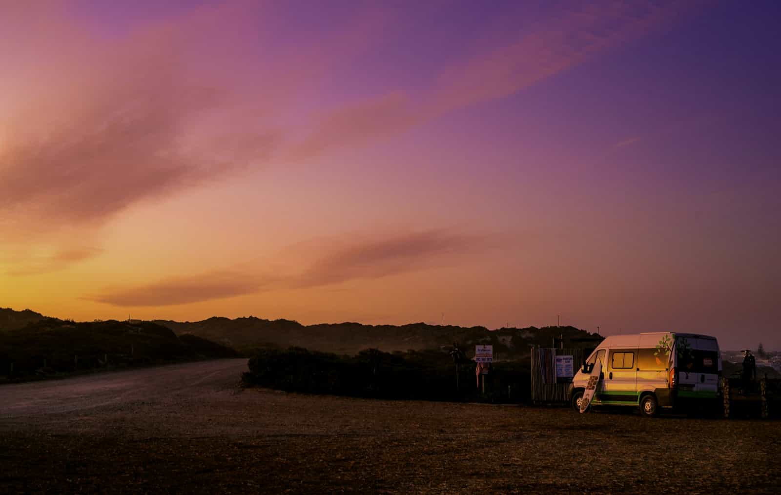 A camper van parked at the end of a dirt road under a pink and orange sunset sky