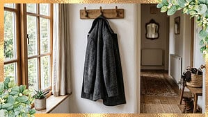 A single wool coat hanging on a wooden hook by a front door entry with natural light from a nearby window and a hallway in the background