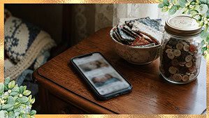 Smartphone on a wooden side table beside a small bowl of folded fabric scraps and a glass jar of buttons