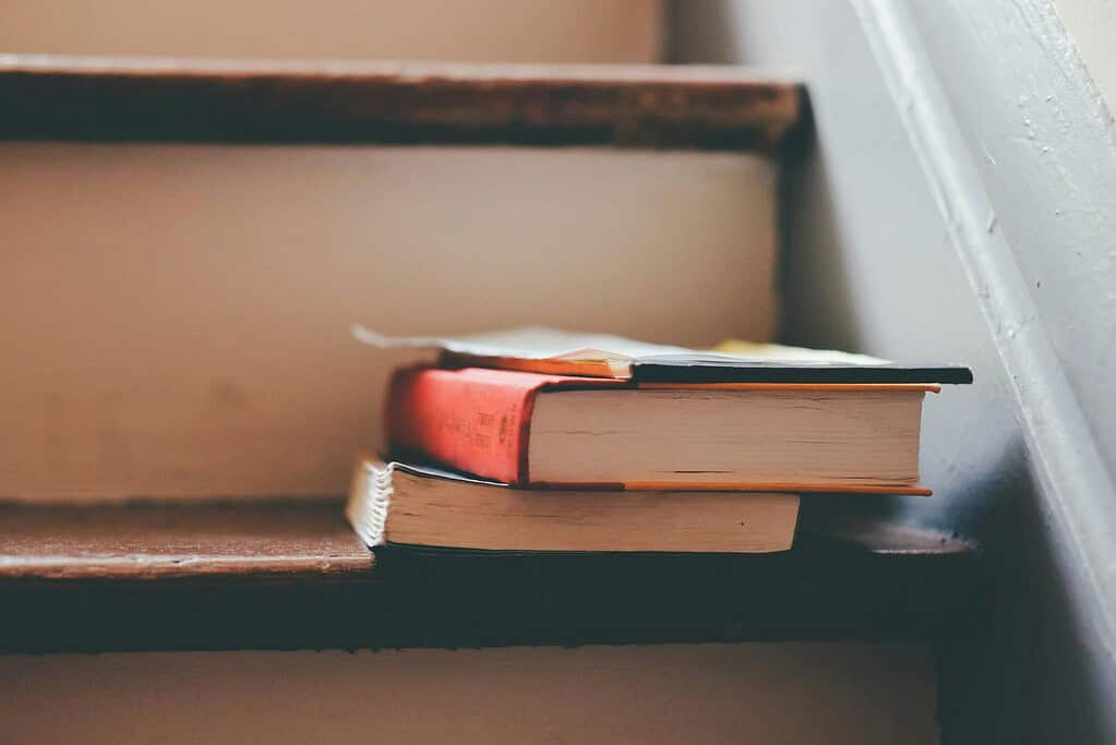 A small stack of hardcover books resting on a wooden staircase step with soft natural light from a nearby window