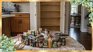 An open empty pantry cabinet in a kitchen with canned goods boxes and jars arranged on a bedsheet on the floor in front of it in bright natural light