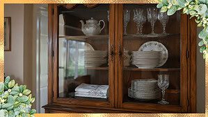 A glass-front cabinet with neatly stacked china plates, a folded tablecloth, and a crystal wine glass all sitting untouched behind closed glass doors