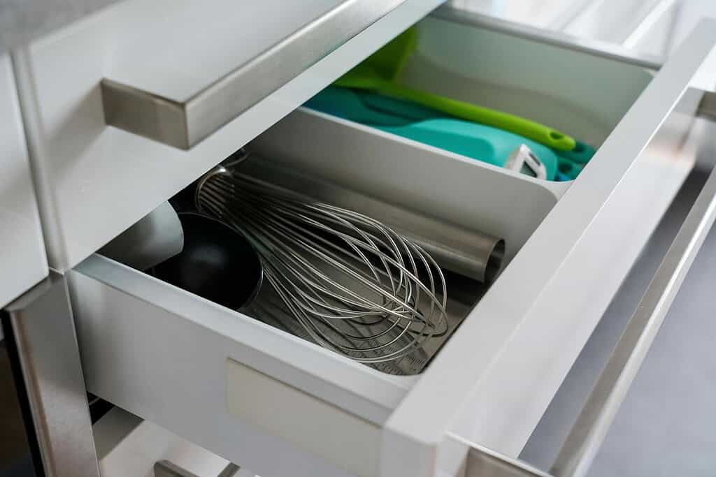 An open wooden kitchen drawer showing a wire whisk and a few cooking utensils neatly arranged inside
