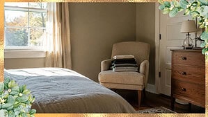 A single upholstered bedroom chair in the corner of a modest American home holding a small stack of folded laundry in soft morning light