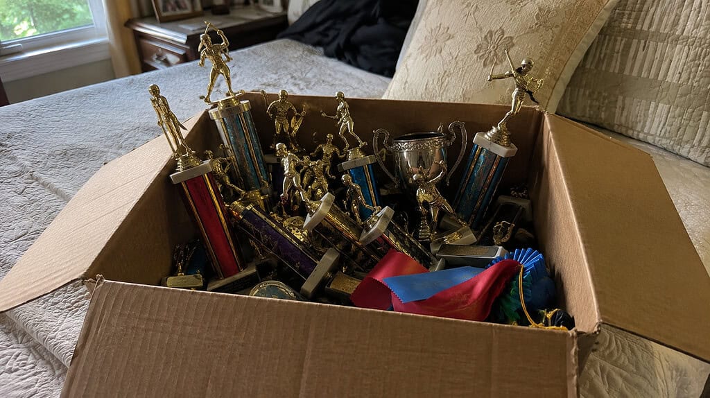 An open cardboard box of a child's old mismatched gold and silver trophies with sports figurines and faded school ribbons sitting on a bed