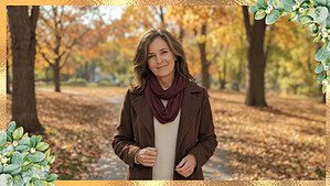 A middle-aged woman with shoulder-length brown hair wearing a chocolate brown wool coat, cream sweater, and burgundy scarf in a sunlit autumn park.