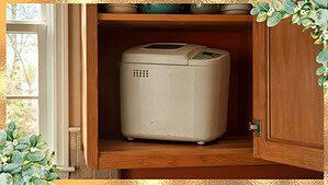 A slightly dusty white bread machine sitting alone on a wooden cabinet shelf against a warm terracotta wall in soft daylight