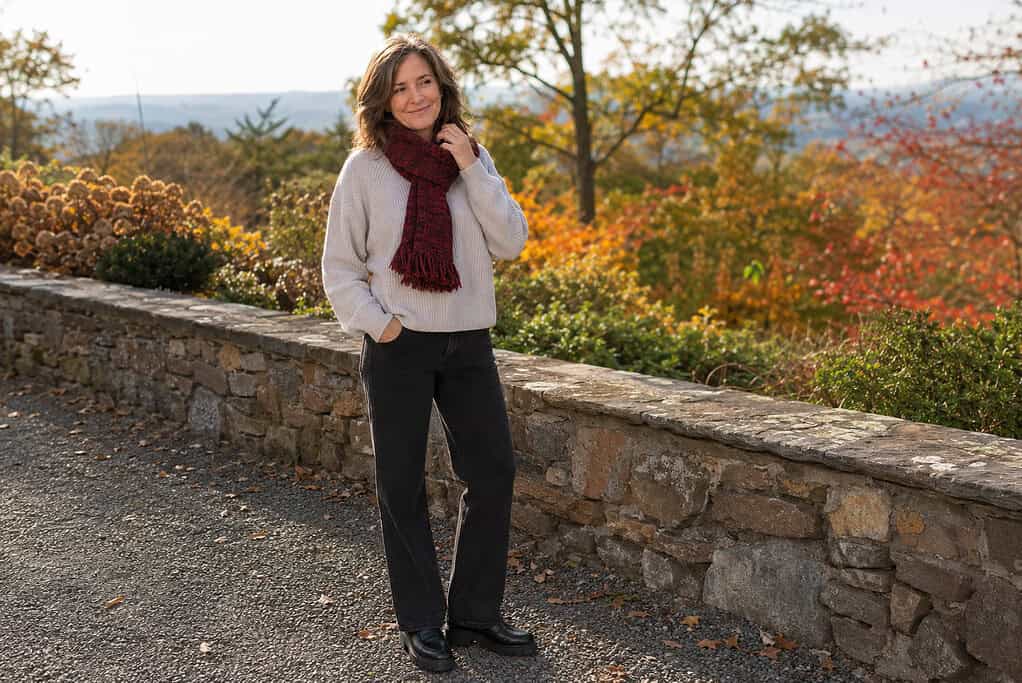 Woman over 50 wearing a burgundy knit scarf, oatmeal sweater, dark jeans, and black loafers beside a stone wall in autumn.