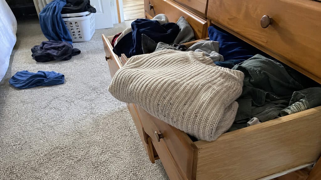 A folded wool sweater on the edge of an open dresser drawer with clothes jumbled around it, the kind of item that's hard to decide whether to keep when decluttering