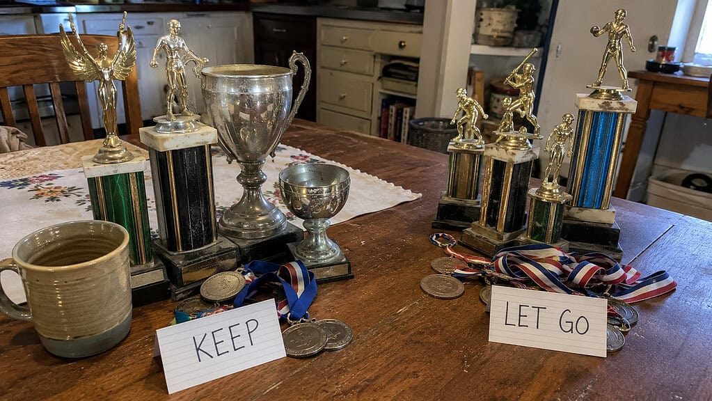Old mismatched gold and silver trophies and medals sorted into two small groups on a kitchen table with a warm mug beside them