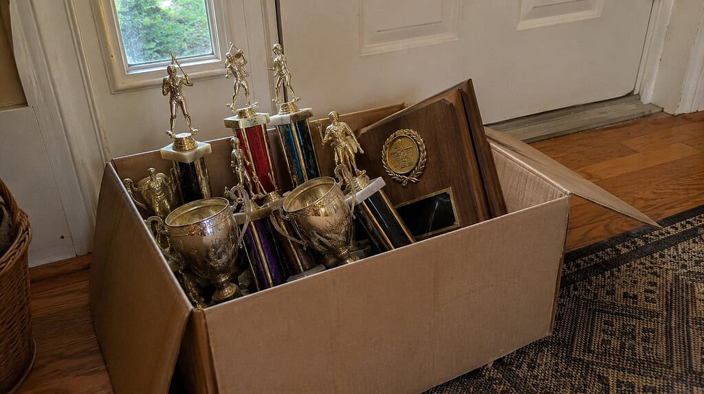 A cardboard box of old mismatched gold and silver trophies and wooden plaques sitting by the front door, ready to be donated