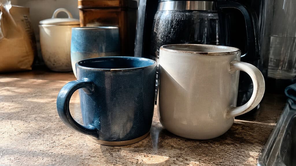 Two well-used favorite coffee mugs, one blue and one larger plain one, sitting together on a kitchen counter near a coffee maker