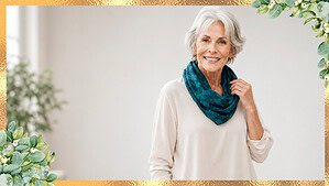 Woman in her seventies with short silver hair wearing an untucked cream overblouse, a deep teal silk scarf, and camel straight-leg trousers, standing in a bright living room