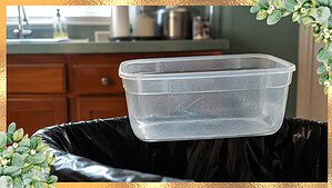 A scratched clear plastic container with no lid on the edge of an open black trash bag, with a sage-green counter and warm wood cabinet behind it in soft daylight