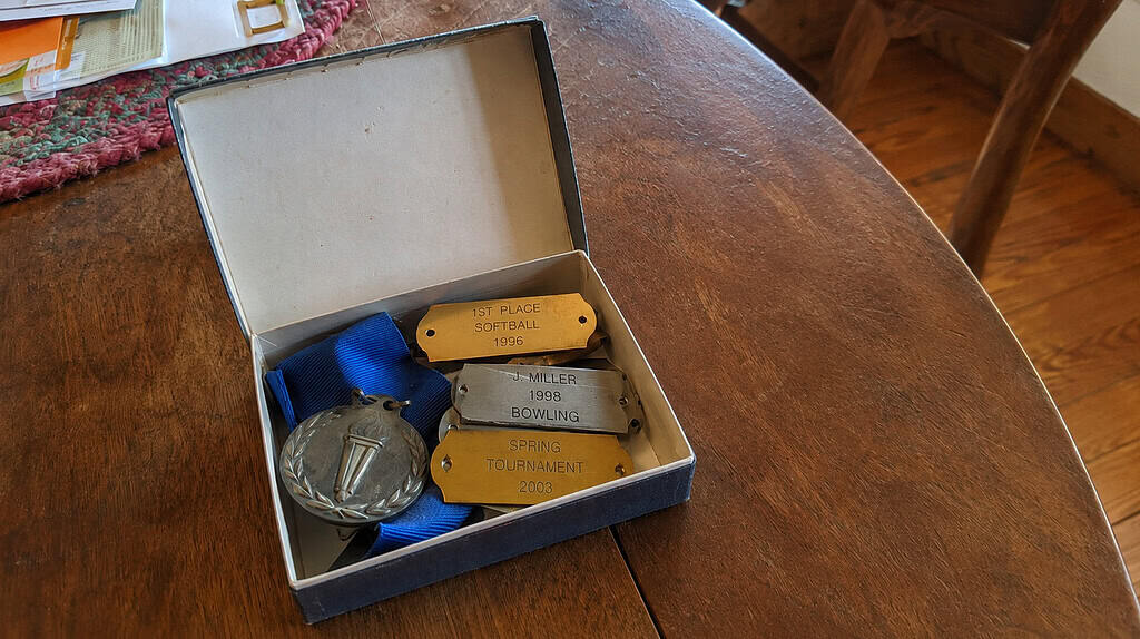 A small open memory box holding a few engraved metal trophy nameplates, a folded blue ribbon, and a flat medal, resting on a table in soft light