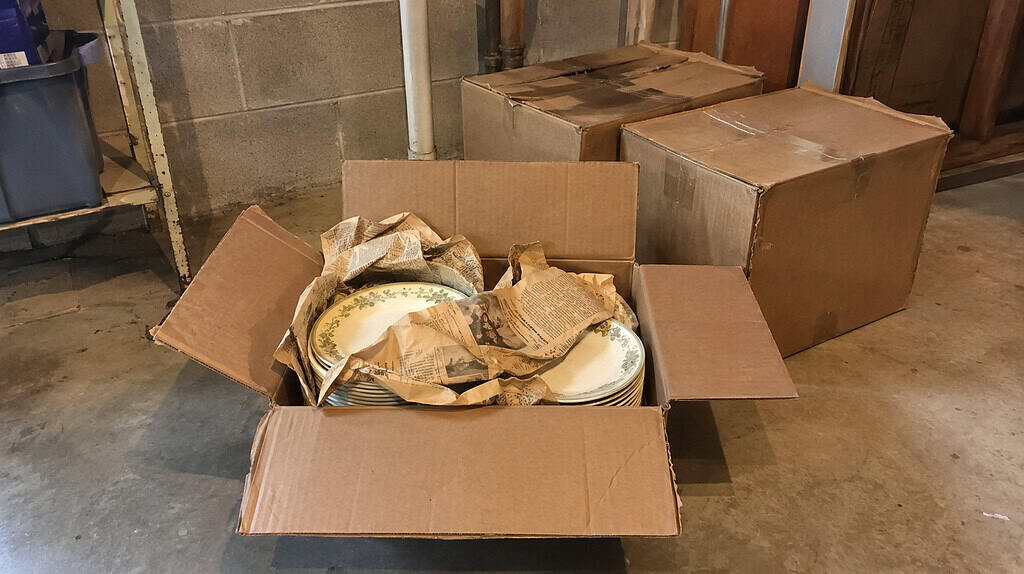 An opened cardboard box on a basement floor revealing stacks of old dinner plates wrapped in yellowed newspaper, with taped boxes behind it