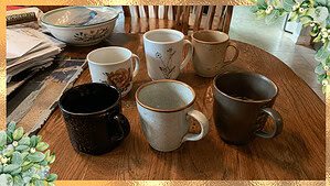 Six mismatched ceramic coffee mugs in different colors and patterns arranged on a worn wooden kitchen table, with a stack of papers and a bowl nearby.