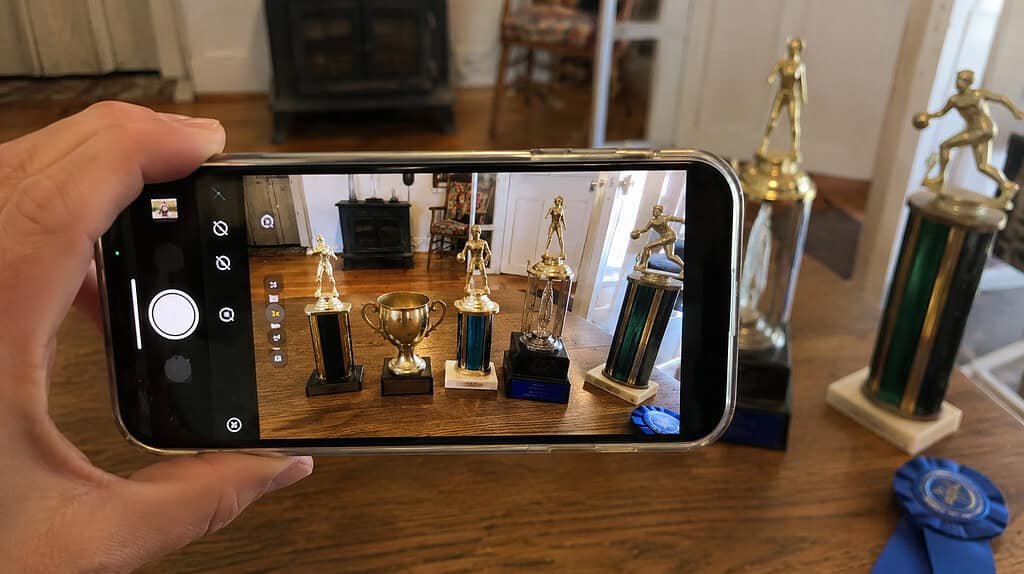A cell phone photographing a row of old mismatched gold and silver trophies and a blue ribbon lined up on a wooden table