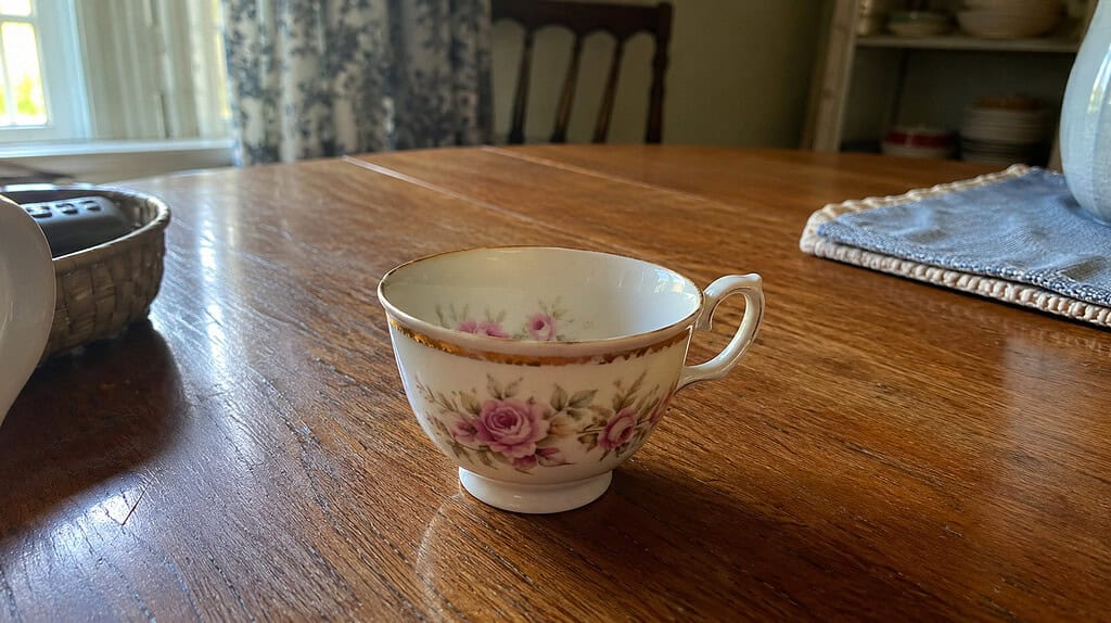 A single old floral teacup with a faded gold rim sitting on a warm wooden kitchen table in soft window light