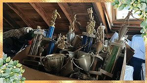 A cardboard box crammed with old mismatched gold and silver trophies of various heights, some with figurines on top and two-handled cups, tarnished and dusty