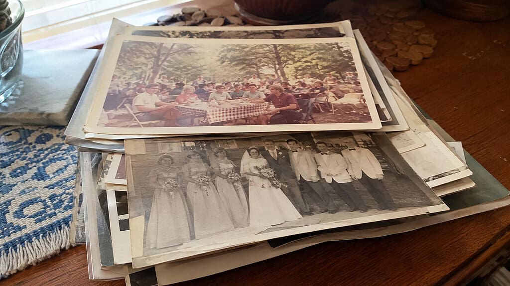 A small pile of very old faded photographs of unknown people, including a group picnic photo and an old wedding party print, on a wooden table