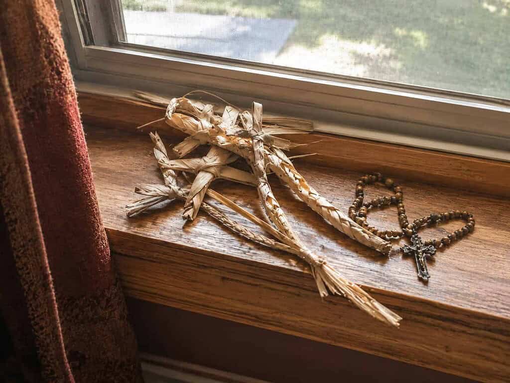 Dried braided palm fronds and a rosary resting together on a wooden windowsill in warm light