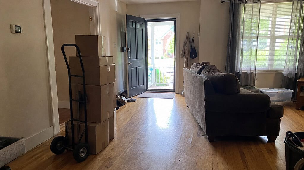 A nearly cleared living room with a hand truck loaded with boxes and a couch pushed toward the open front door, ready for pickup