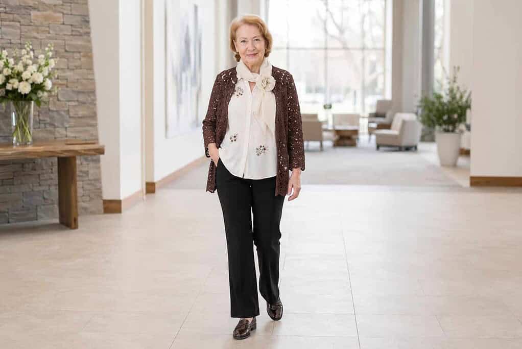 Woman in her seventies with short blonde hair wearing an embroidered white blouse, a brown lace cardigan, black trousers, and brown loafers in a bright modern church lobby.