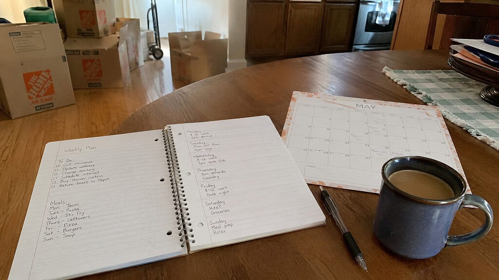 An open spiral notebook with a handwritten weekly plan and a wall calendar on a kitchen table with a pen and mug, boxes in the background