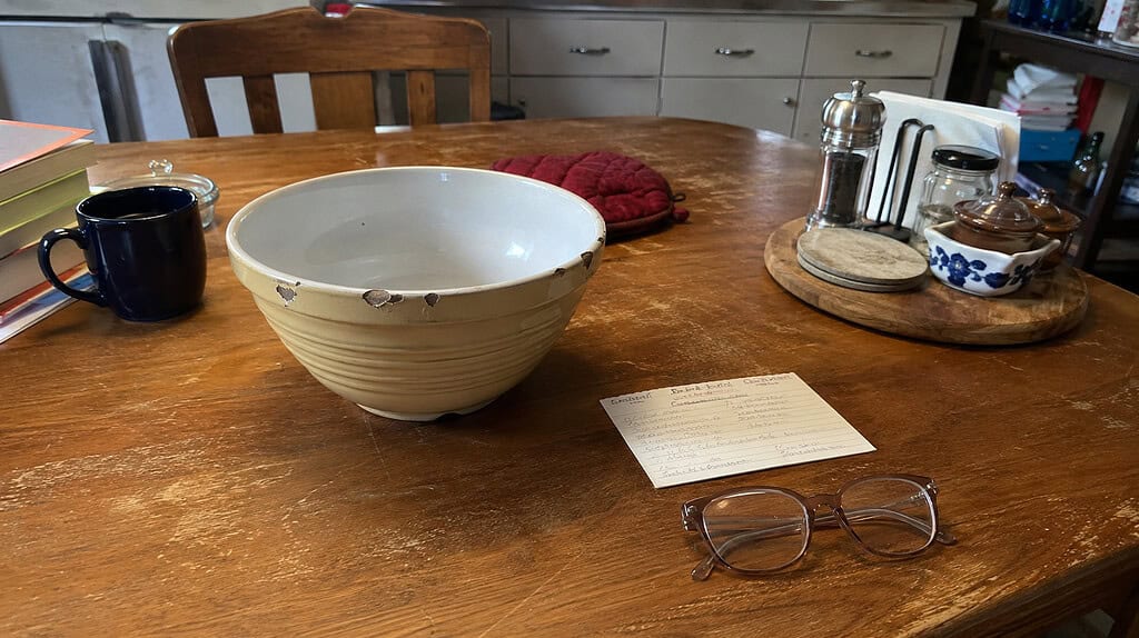 A worn kitchen table holding an older parent's everyday things, a chipped mixing bowl, a handwritten recipe card, and reading glasses