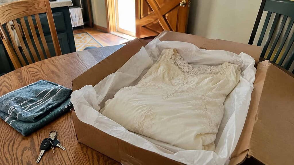 An ivory old wedding dress folded into a tissue-lined cardboard box on a kitchen table, ready to donate