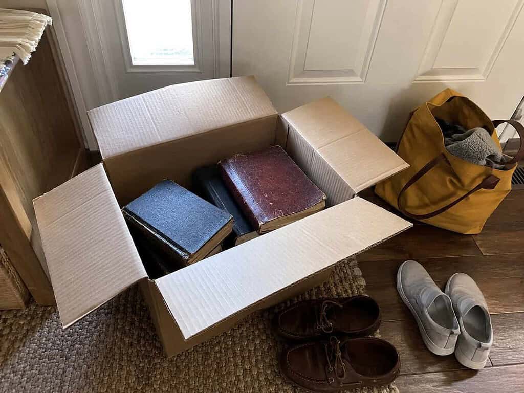 An open cardboard donation box holding a few old Bibles and books on the floor near a front door