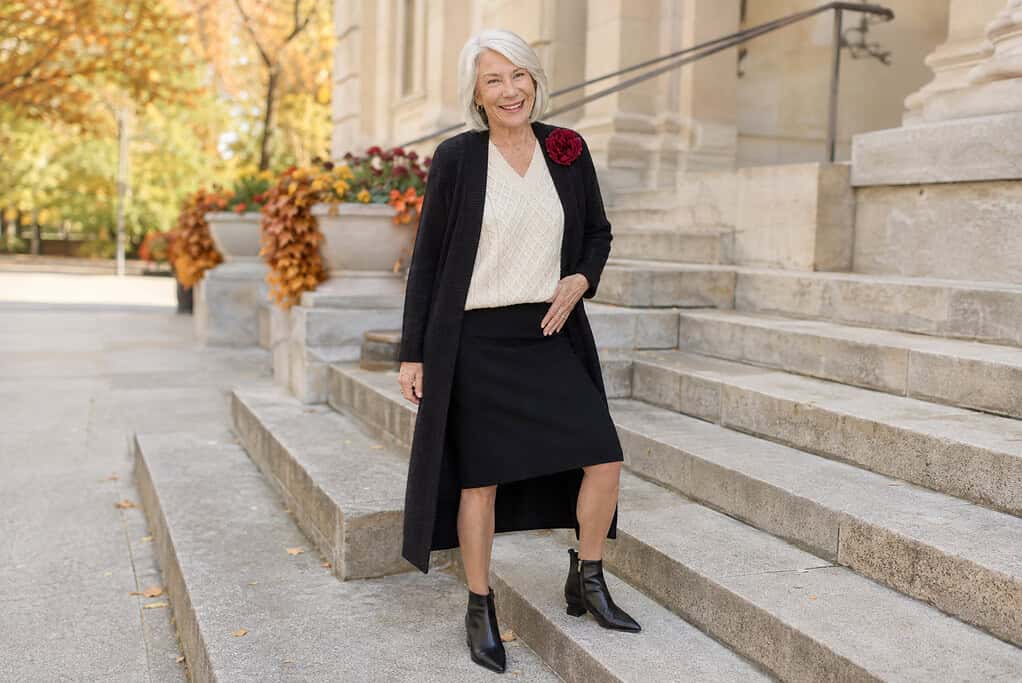 Confident older woman wearing a cream V-neck sweater, long black cardigan, black midi skirt, pointed ankle boots, and burgundy flower brooch for fall.