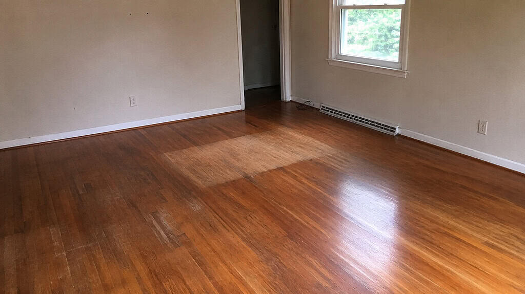 An empty cleared-out living room with a bare floor showing one brighter square where a chair once sat, soft daylight through an uncovered window