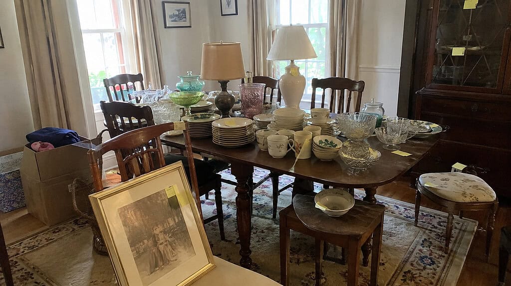 A dining room set up for an estate sale with dishes, glassware, lamps, and small furniture arranged with price stickers and a donation box nearby