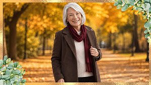 Confident older woman wearing a chocolate brown coat, cream knit sweater, burgundy scarf, and dark trousers in a golden autumn park.