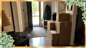 A front hallway in a home partway through being cleared, with stacked moving boxes, a coat hanging by the door, and house keys on a side table