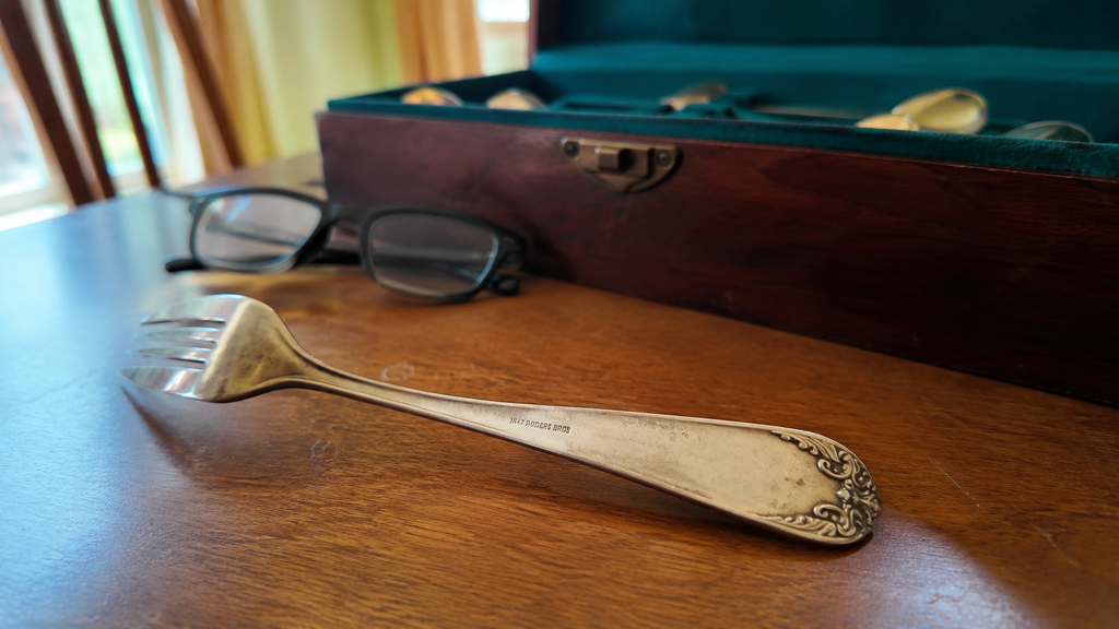 The back of an old silver fork handle showing a small hallmark stamp, next to reading glasses and an inherited silverware chest