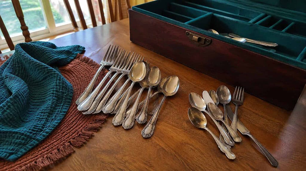 Old inherited silverware sorted into two piles on a kitchen table beside an open silverware chest