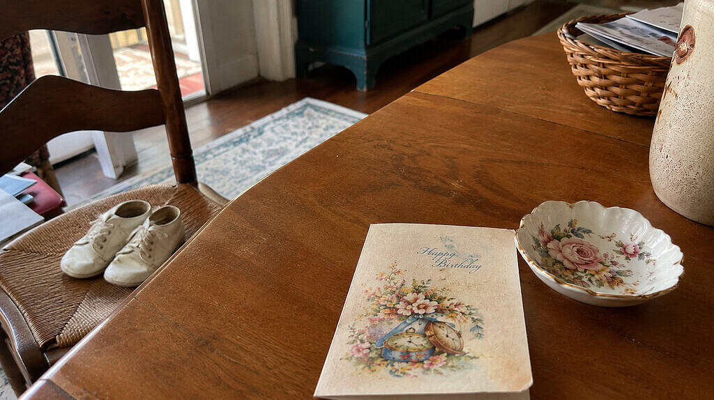 A few cherished keepsakes on a wooden table, an old birthday card, a pair of baby shoes, and a small inherited floral dish, in soft daylight