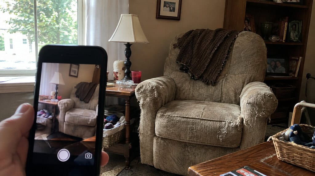 A cell phone held up taking a picture of an old worn armchair in the corner of a living room in soft daylight