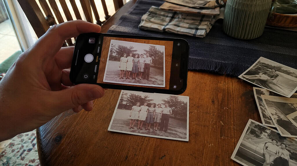 A cell phone held over an old vintage family photograph laid flat on a table, taking a digital copy of the print, with more old photos beside it